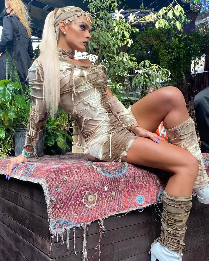 Femme trans aux cheveux blancs, en robe jaune, posant sur un canapé blanc.