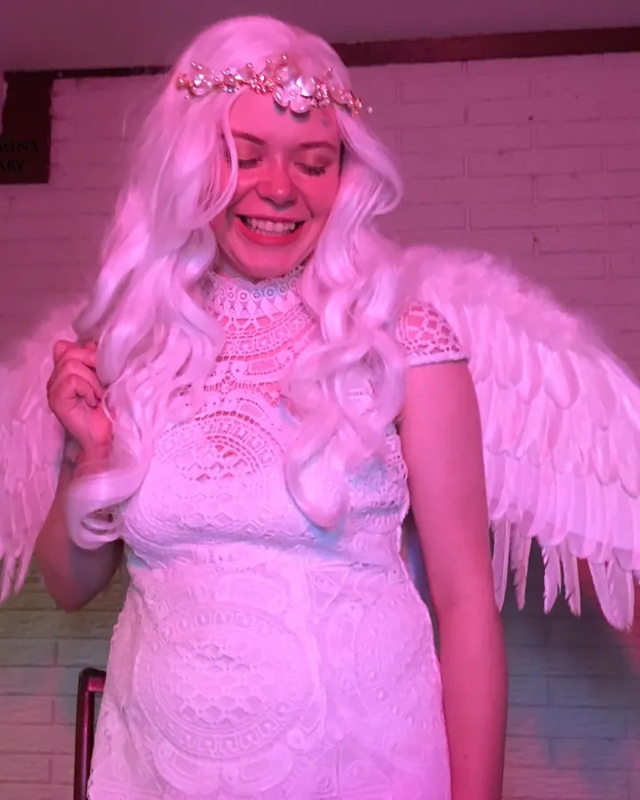 Femme trans aux cheveux platine, en robe blanche dentelle, ailes d'ange, couronne de fleurs, souriant sous lumière rose.