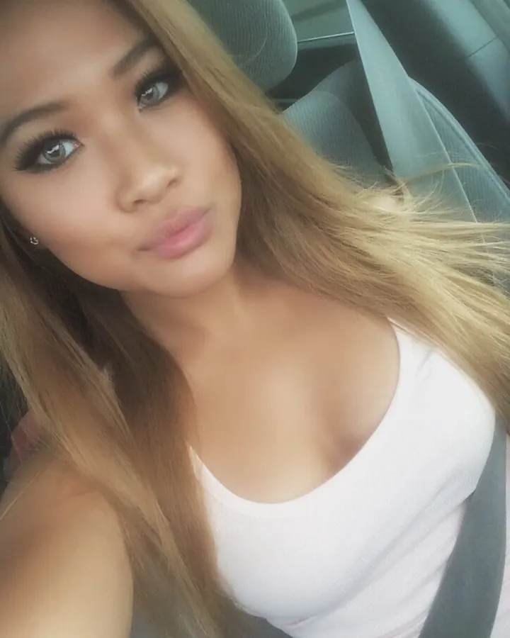 Femme trans aux cheveux blonds, en haut blanc, portant une casquette, assise dans une voiture, regardant vers l'appareil.