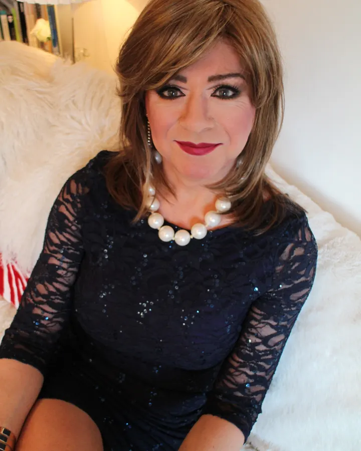 Femme trans aux cheveux châtains, en robe bleue à dentelle, assise sur un canapé blanc avec un collier de perles.