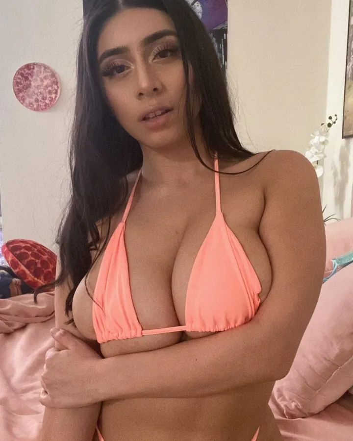Femme trans asiatique aux cheveux bruns, portant un bikini orange, assise sur un lit rose, tenant ses seins avec ses bras croisés.