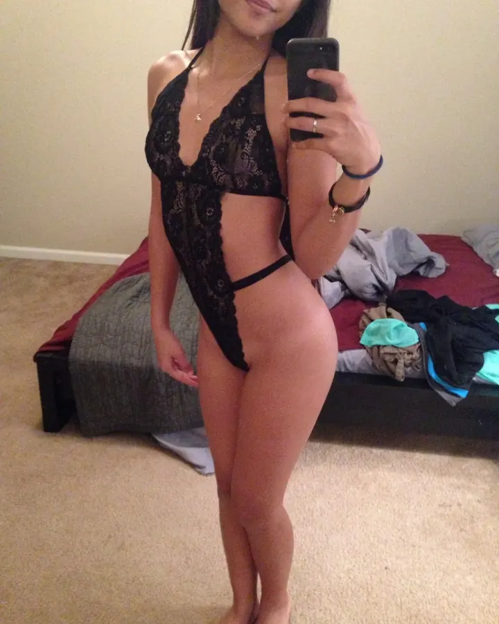 Femme trans asiatique aux cheveux bruns, portant un haut noir, se tenant debout dans une chambre, tenant un téléphone pour prendre un selfie.