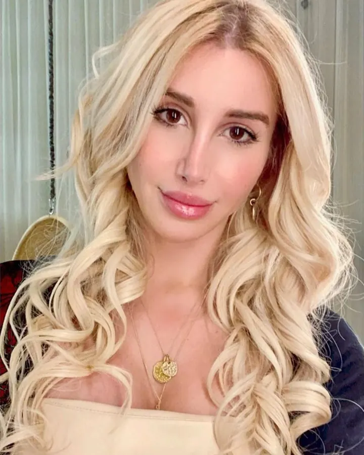 Femme trans européenne aux cheveux blonds, portant un haut beige, assise près d'une porte blanche, tenant une sucette dans sa bouche, regardant de côté.