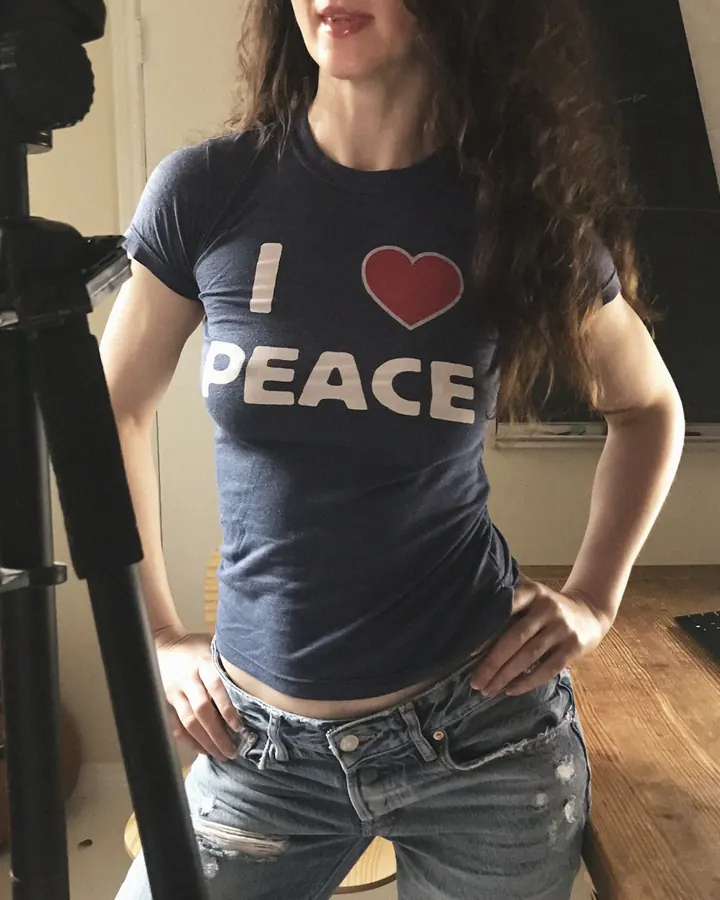 Femme trans en tenue décontractée, t-shirt bleu "I ♥ PEACE", jeans déchirés, pose devant un trépied.
