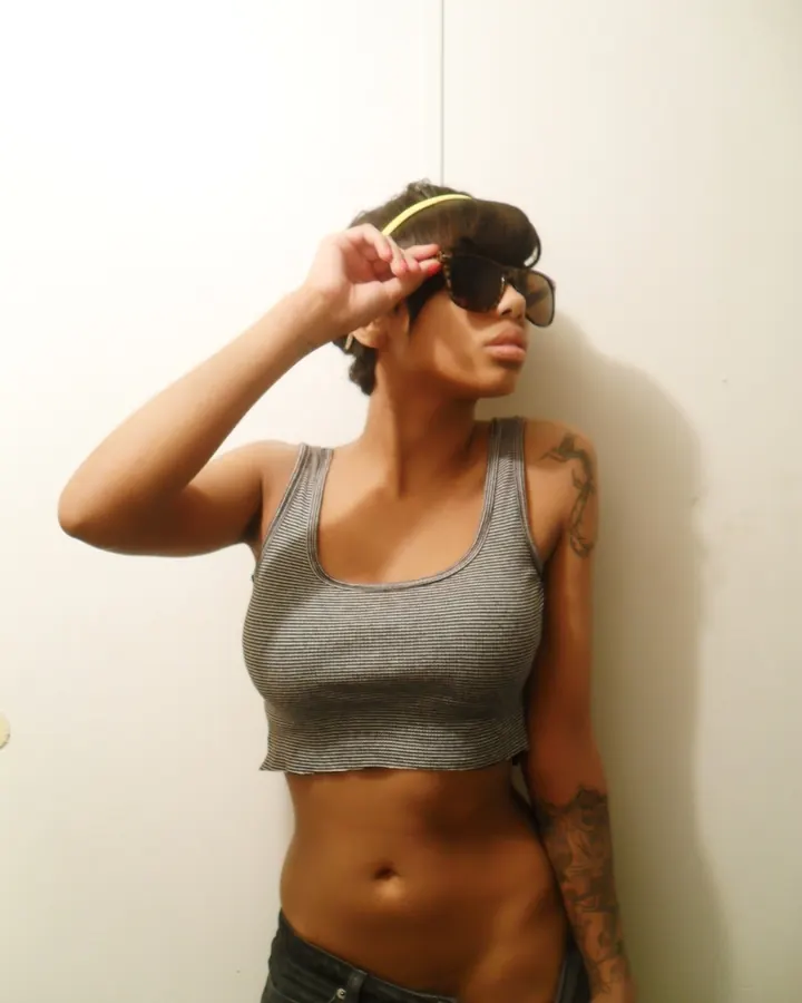 Femme trans aux cheveux bruns, torse nu, posant sur un canapé beige, avec une main sur son entrejambe.