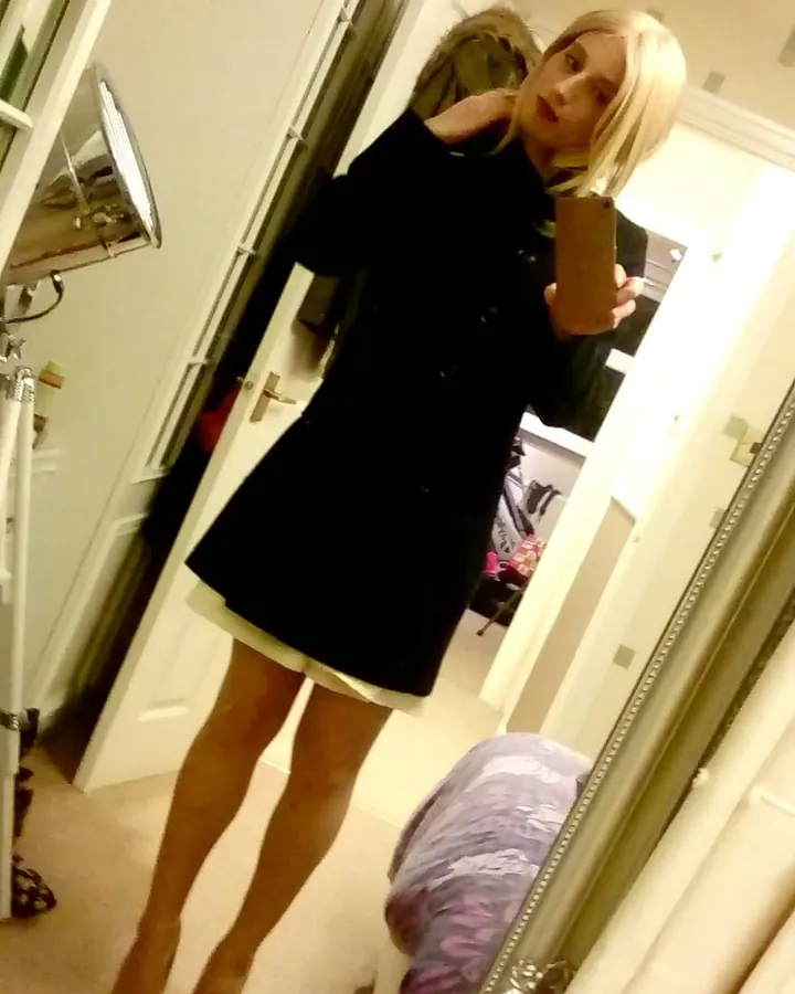 Femme trans aux cheveux blonds, portant une chemise blanche et une jupe noire, posant devant une porte vitrée avec des volets.