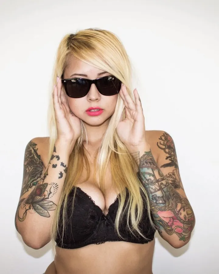 Femme trans aux cheveux blonds, tatouages sur les bras, portant un soutien-gorge noir et des lunettes de soleil, posant devant un mur blanc.