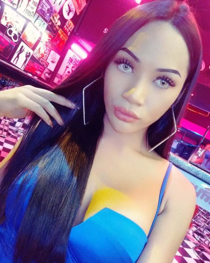 Femme trans aux longs cheveux noirs, en tenue bleue, posant dans un bar aux lumières roses.