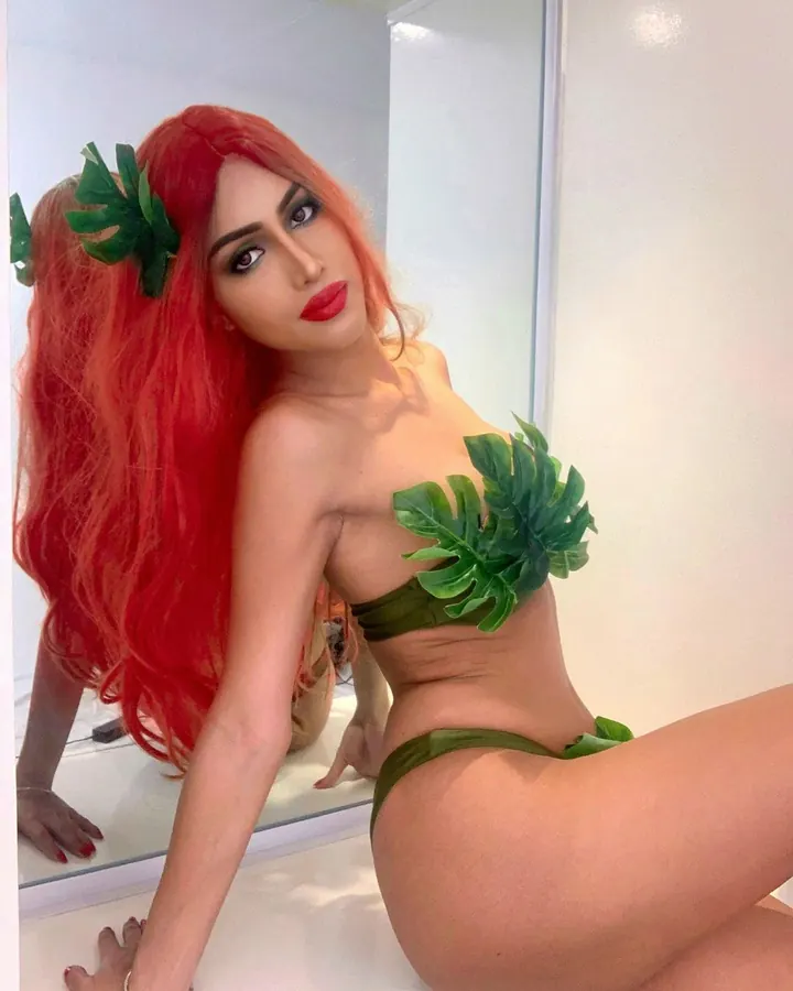 Femme trans aux cheveux roux portant un haut vert avec des feuilles, posant devant un miroir.