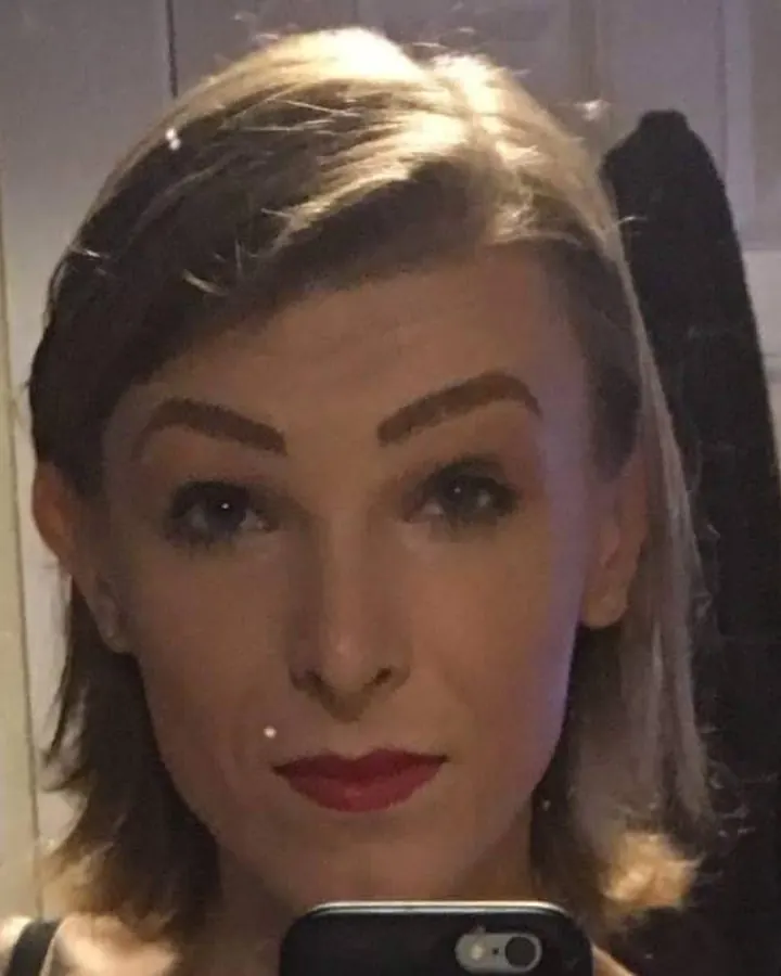 Femme trans aux cheveux blonds portant un haut noir, assise sur un lit, les jambes croisées.