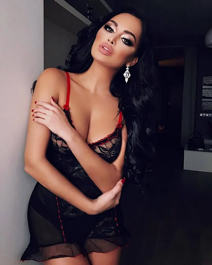 Femme trans aux cheveux noirs portant un haut noir, posant près d'une table de cuisine.
