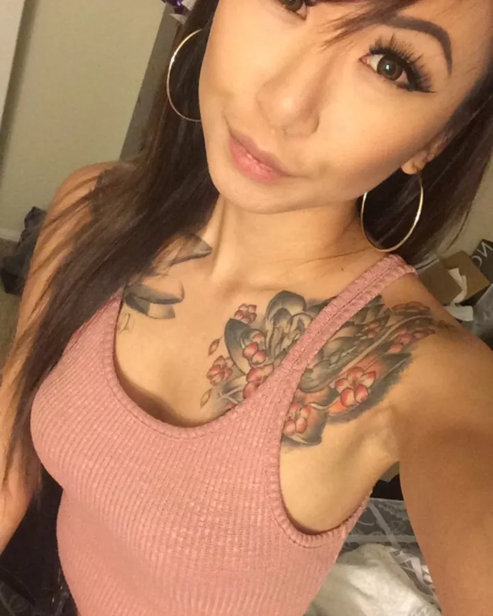 Femme trans aux cheveux noirs, portant un haut rose pâle, avec un tatouage floral visible sur la poitrine, prenant un selfie.